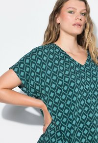 Blusa verde a fantasia con scollatura a V, maniche corte e una vestibilità ampia, realizzata in tessuto leggero con un design geometrico.