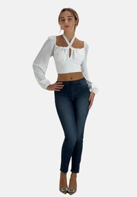 Hvid cropped top med pufærmer og en front nøglehul, parret med mørke skinny jeans og glitrende beige hæle. Ensfarvet baggrund.