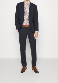 Traje navy con una camisa de patrón floral, cinturón de cuero marrón y zapatos de vestir marrones. El traje tiene un corte entallado y una textura suave.