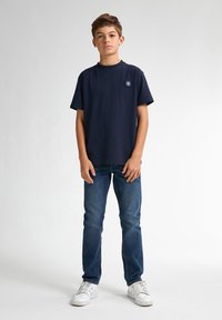 Navy katoenen t-shirt met korte mouwen en een klein logo op de borst, gecombineerd met blauwe spijkerbroeken en witte sneakers. Beide passen comfortabel.
