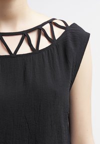 Top sin mangas negro con un tejido texturizado, que presenta tiras en forma de cruz a lo largo del escote para un detalle adicional.