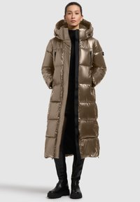 Long, padded coat in shiny taupe nylon with a hood, featuring side zip pockets and a zip front closure. Paired with knee-high black boots.