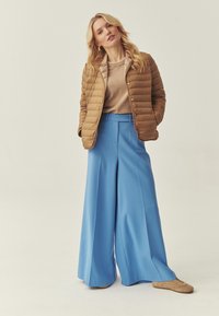 Femme blonde portant une veste matelassée beige, un pull beige, un pantalon bleu à jambes larges et des baskets beiges, debout devant un fond uni.