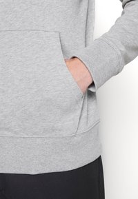 Mano parzialmente dentro della tasca anteriore di una felpa grigia chiara, che mostra il polsino a coste e parte dei pantaloni neri.