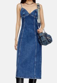 Kvinna iklädd en åtsittande denim-middklänning med tunna band, som håller en sliten denimaxelväska med en grå rem.