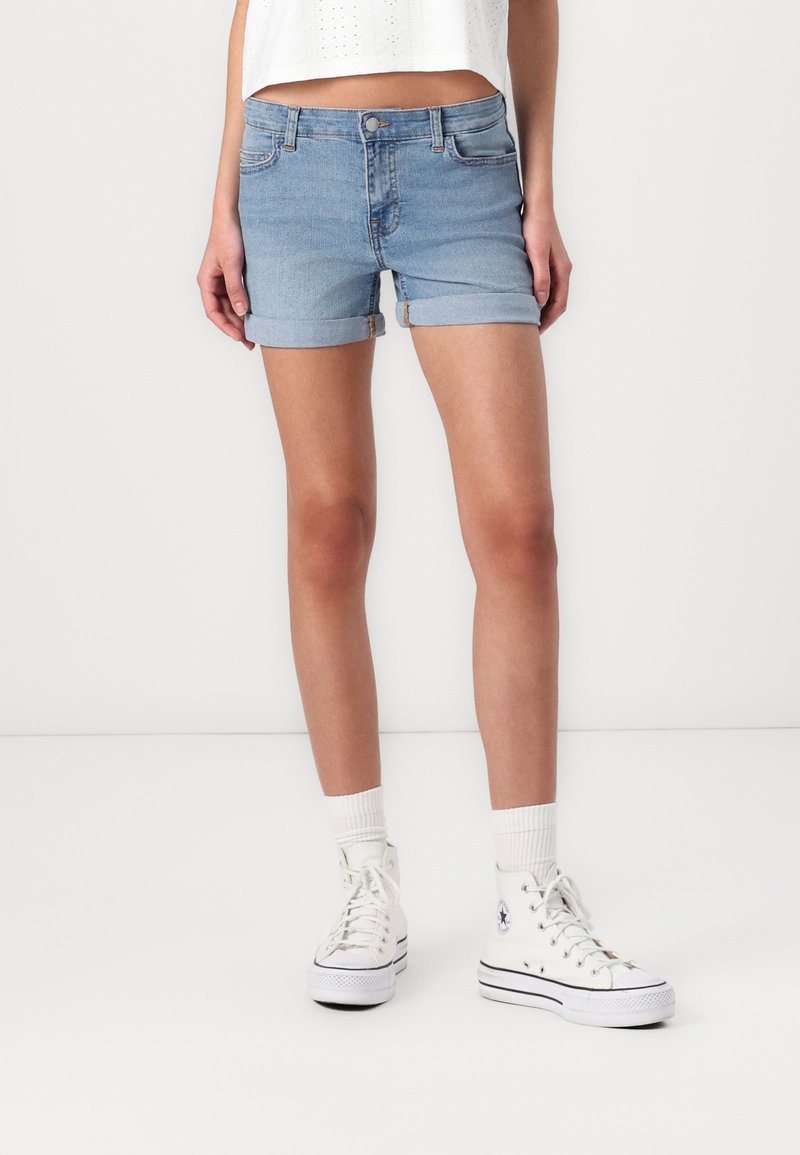 Ljusblå denimshorts med uppvikta fållar, femfickorsdesign och knäppning med knapp. Bärs tillsammans med vita högklackade sneakers och längdstrumpor.