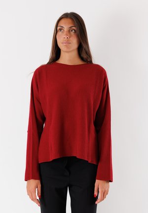 Donna in piedi contro uno sfondo bianco, che indossa un maglione rosso a coste largo e pantaloni neri, guardando leggermente verso l'alto e di lato.