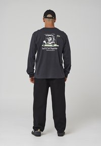 Camiseta negra de manga larga con gráfico en la espalda de un esqueleto en un carrito de golf, con el texto "Departamento Surf & Turf", combinada con pantalones oscuros.