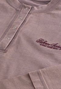 Pikend varrukatega pluus mauve kangast, ümarate kaeluste ja viie nupuga, millel on tume burgundi värvi tikandiga logo.