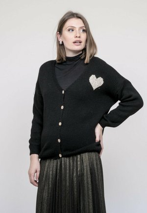 Femme portant un cardigan noir boutonné avec un cœur blanc sur la poitrine, un col roulé noir et une jupe plissée métallique, posant avec la main sur la hanche.