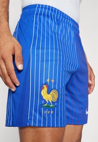 Blå sportshorts med vertikala vita ränder, med en gul broderad tuppemblem och två stjärnor ovanför. Nike-logotypen är närvarande.