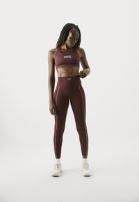 Sieviete, kas ģērbjas bordo krāsas Nike sporta krūšturī un legingos, pielāgo jostu, stāvot pret baltu fonu sporta apavos.