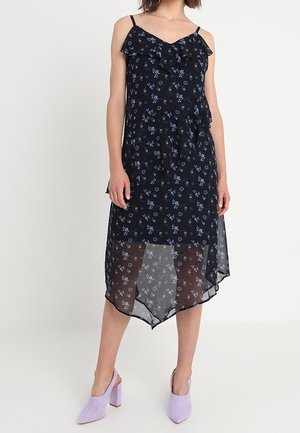 Frau trägt ein schwarzes ärmelloses Blumenkleid in Midilänge mit Rüschen und transparentem asymmetrischem Saum, kombiniert mit lavendelfarbenen Slingback-Schuhen mit Blockabsatz.