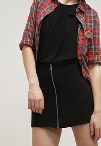 Mini gonna nera con zip frontale, dotata di due tasche laterali. Abbinata a una camicia a quadri rossa, che presenta un look casual a strati.