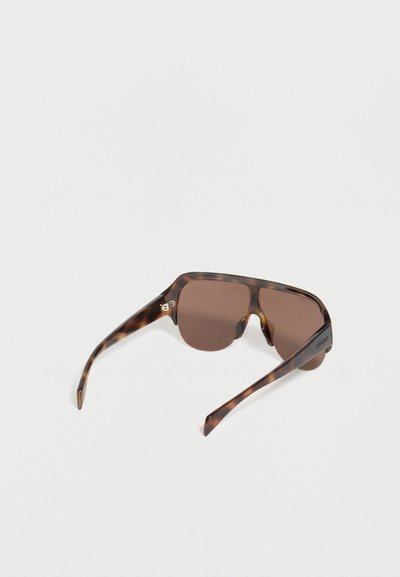 Lunettes de soleil rectangulaires surdimensionnées teintées marron avec une monture écaille de tortue et le logo "Gucci" sur la branche gauche, présentées en angle sur une surface blanche.