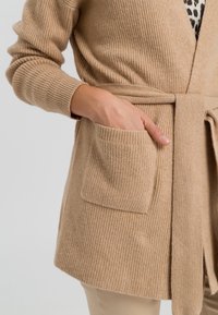 Kvindes hånd placeret i lommen på en beige ribbet strik cardigan, der er bundet i taljen over en leopardmønstret top og lyse bukser.