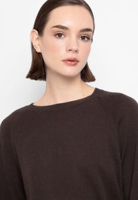 Maglione marrone a maglia con scollatura ampia e maniche raglan, caratterizzato da una texture morbida e una finitura liscia. Appare rilassato e informale.