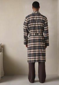 Kariertes Bademantel in Dunkelblau, Beige und Braun, mit Kragen, langen Ärmeln und einem Taillenband; kombiniert mit brauner Hose. Schlichter Hintergrund.