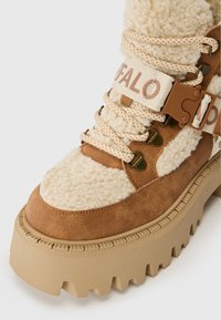 Bota plataforma marrón y beige con suela gruesa, forro de felpa, cordones, y una correa de tela con hebilla de plástico y texto "BUFFALO".