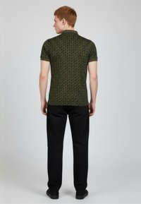 Olivově zelené polo triko s krátkými rukávy a opakujícím se vzorem. Má límec a kontrastní lemování. Nošeno s černými kalhotami a botami.