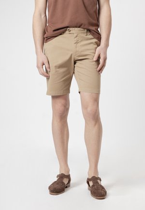 Hombre que lleva pantalones cortos beige hasta la rodilla, camiseta marrón y sandalias de cuero trenzado marrón, de pie contra un fondo claro y sencillo.