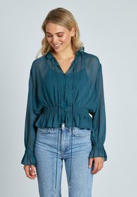 Blusa lunga a maniche trasparenti color teal con vita regolabile, bottoni sul davanti e polsini arricciati. Indossata con jeans blu.