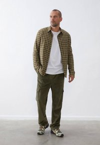 Olive green checkered shirt, white crew-neck t-shirt, olive corduroy pants, and camo patterned shoes. Smooth, casual fit, minimal accessories.
