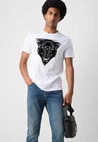 Camiseta blanca de algodón con un gráfico de pantera negra y ojos verdes. Combinada con unos vaqueros de mezclilla azules y un bolso negro de hombro.