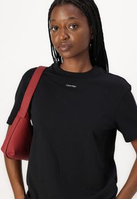 Sort bomulds t-shirt med korte ærmer, der har et lille hvidt "Calvin Klein" logo på brystet. Holder en rød lædertaske.