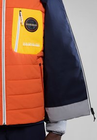 Chaqueta acolchada con un cuerpo naranja, mangas azul marino y un bolsillo en el pecho amarillo con un logotipo redondo y texto. Cierre con cremallera y puños.