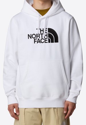 Hvid hættetrøje med sort "The North Face"-logo på brystet, båret over en sennepsskjorte, parret med lyse beige bukser.