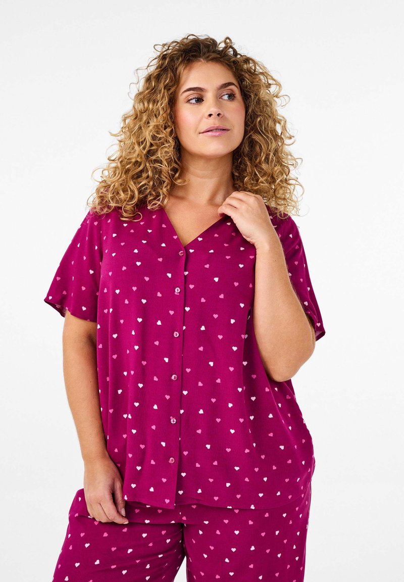 Magenta pyjamas med korta ärmar, med V-halsringning och knappfront. Tyget har ett hjärtmönster i nyanser av rosa och vitt.