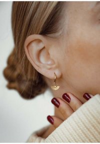 Pendiente de aro dorado con un charm en forma de abanico, que presenta una superficie texturizada. El modelo tiene el cabello rizado y las uñas pintadas de color burdeos.