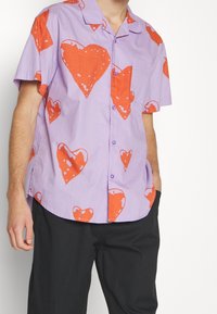 Košile s krátkým rukávem z světlé fialové látky s velkými červenými srdci. Zapínání na knoflíky vpředu s límečkem a jednou kapsou na prsou.
