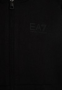 Felpa nera con zip realizzata in cotone con tessuto strutturato. Presenta un discreto logo EA7 Emporio Armani in rilievo sul petto.