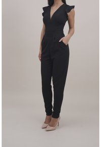 Svart jumpsuit med volangärmar, djup urringning, figursydd midja och sidofickor. Bärs med nakna högklackade skor. Slät textur.