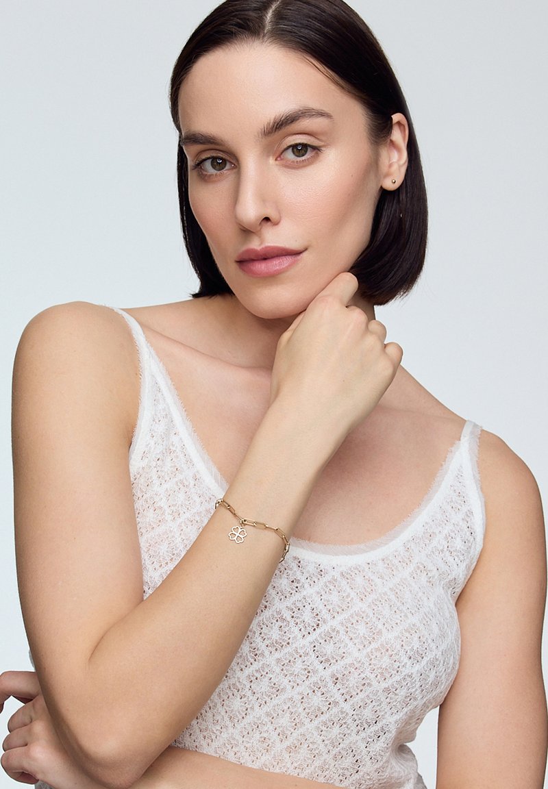 Złota bransoletka z kwiatowym wisiorkiem, fakturowanym łańcuchem i ogniwami. Noszona na nadgarstku, w parze z białą, przezroczystą bluzką na ramiączkach. Widać gładką karnację.