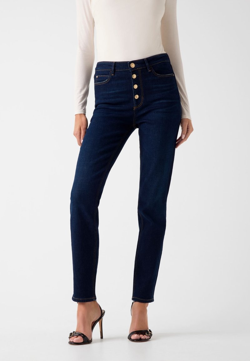 Donkere denim jeans met hoge taille, knoopsluiting, vijf zakken en subtiele contrasterende stiksels. Strakke pasvorm met enkel lengte.