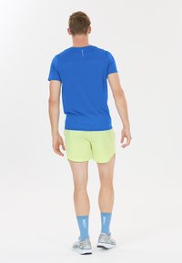 Mand iført en blå kortærmet skjorte, lysegrønne shorts, blå sokker og grå atletiksko, stående med ryggen mod på en hvid baggrund.