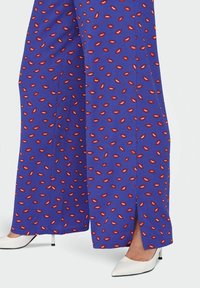 Pantalones de pierna ancha en un vibrante tejido púrpura, con un patrón de labios rojos, con aberturas laterales y combinados con tacones blancos de punta. 