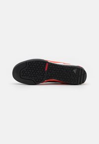 Semelle en caoutchouc noir avec motif géométrique de la bande de roulement et étiquette rouge "TRAXION", vue partielle d'une basket Adidas blanche et rouge au-dessus.
