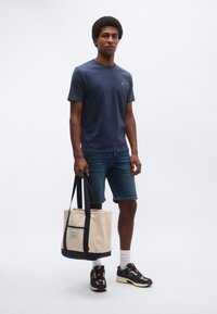 Granatowa koszula z krótkim rękawem, dżinsowe szorty oraz wzorzyste tenisówki. Trzyma beżową bawełnianą torbę na ramię z ciemnoniebieskimi akcentami i metką.