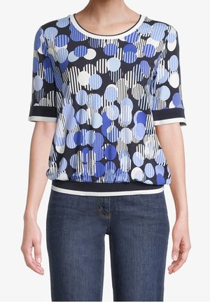 Kobieta w krótkim rękawie bluzce w niebieskie i białe kropki oraz paski, w ciemnoniebieskich dżinsach, stoi z rozluźnionymi ramionami.