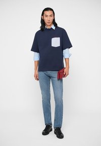 Námornícky veľký tričko s svetlomodrou kapsou, vrstvené cez svetlomodrú košeľu, spárované so svetlými džínsami a čiernymi teniskami. Červený doplnok.