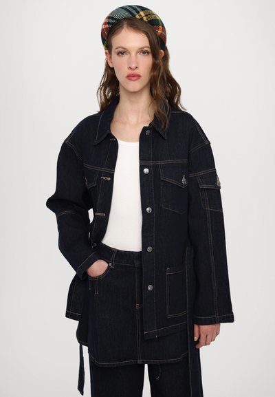 Mørkeblå denimjakke med store lommer, oransje søm og trykknapper, kombinert med en hvit topp og mørke denimbukser.