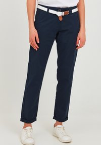 Granatowe spodnie z bawełny z plecionym białym paskiem z brązowym akcentem. Nogawki z mankietami i slim fit. Widoczne białe sneakersy.