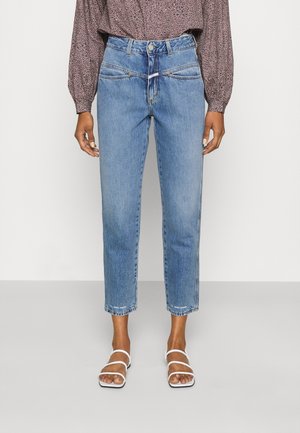 Persoon die een lichtblauwe cropped high-waist jeans draagt met een detail aan het voorpaneel, een roze blouse met lange mouwen en een patroon, en witte sandalen met bandjes.