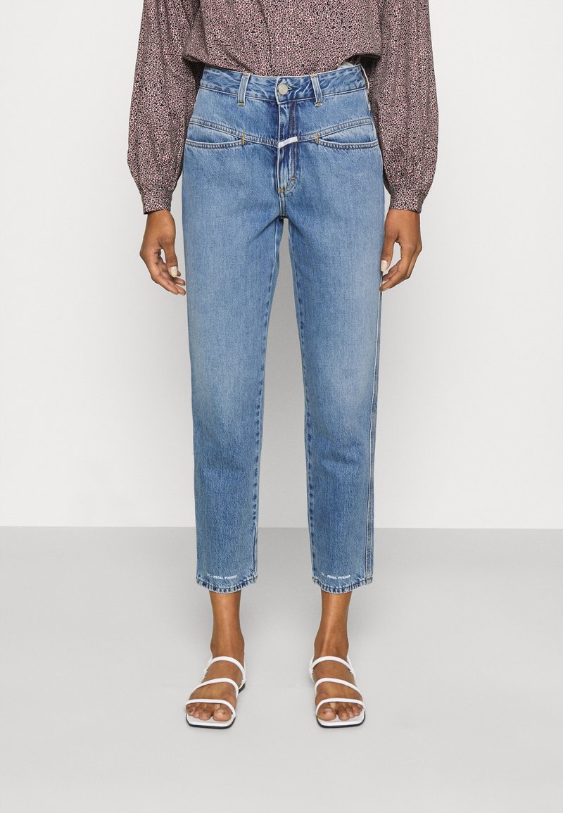Personne portant un jean court taille haute bleu clair avec un détail sur le panneau avant, un chemisier à manches longues rose à motifs et des sandales blanches à lanières.