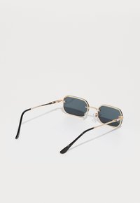 Óculos de sol de metal dourado com lentes escuras octagonais, hastes de plástico preto e almofadas de nariz transparentes. Finos detalhes dourados nas dobradiças.