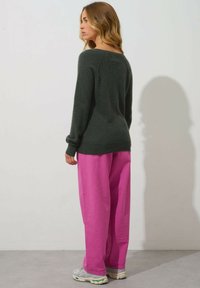 Dunkelgrüner Strickpullover mit langen Ärmeln und weitem Halsausschnitt, kombiniert mit locker sitzenden, leuchtend rosa Ripp-Hosen. Neutrale Sneaker runden den Look ab.